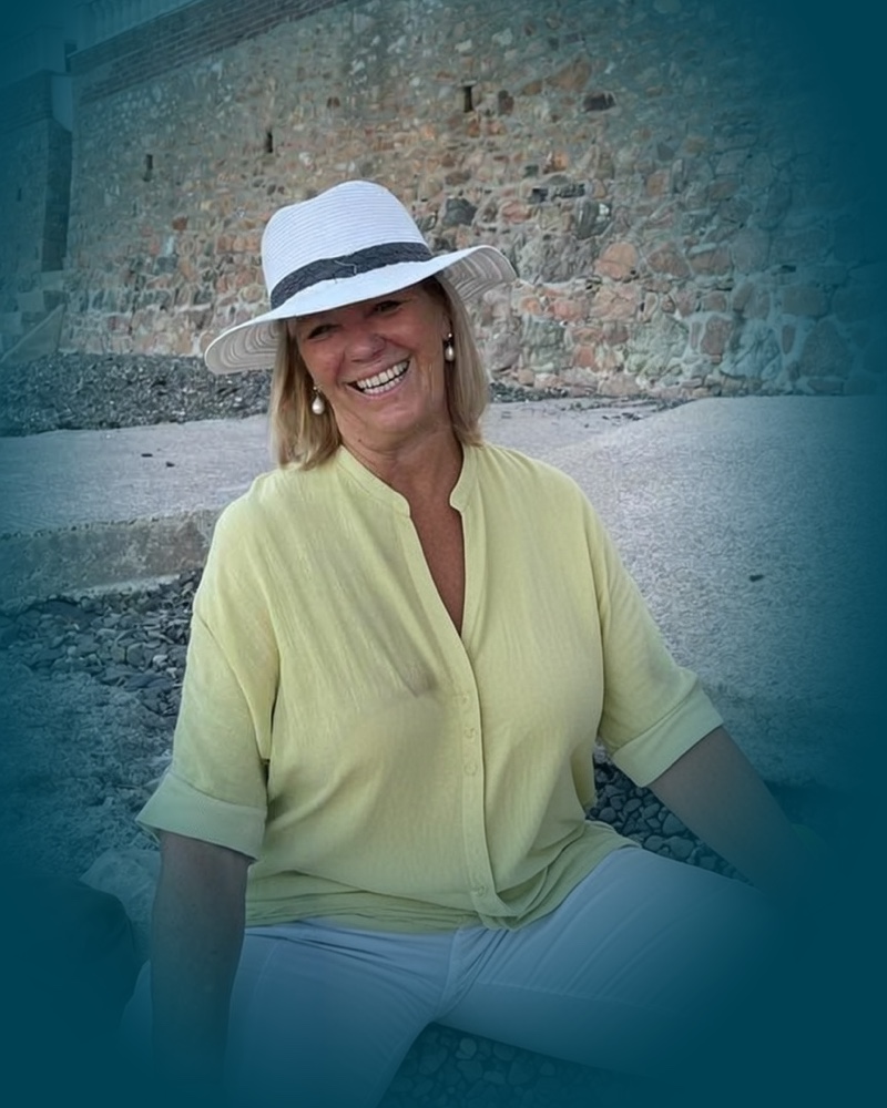 Patricia Reay, sitting on a pebble beach in a white sun hat